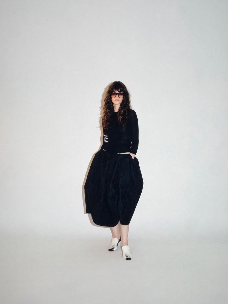 Model wearing dark sunglasses, a black long-sleeve top, a voluminous black bubble skirt, and white heels on a white studio background.