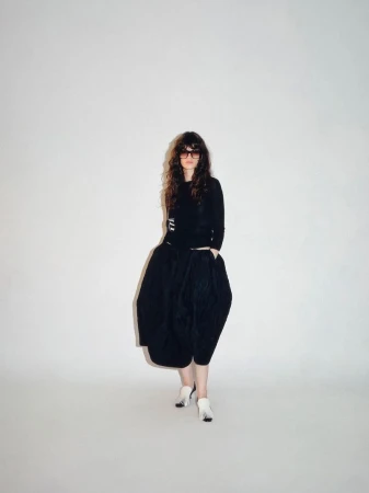 Model wearing dark sunglasses, a black long-sleeve top, a voluminous black bubble skirt, and white heels on a white studio background.