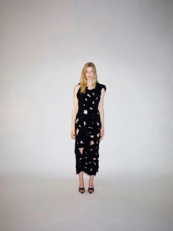 Fashion model wearing a textured black cutout midi dress and black strappy heels against a neutral studio backdrop.