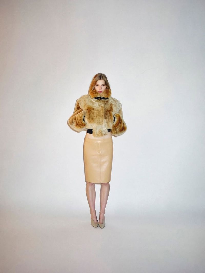 Model posing in a cropped tan fur jacket and beige leather midi skirt against a plain studio backdrop.