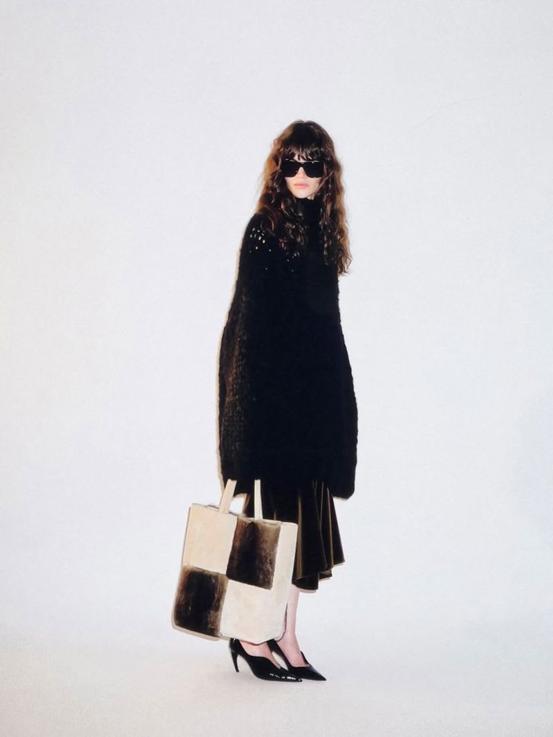 Fashion model wearing an oversized black knit top, dark sunglasses, and holding a checkered shearling tote bag against a white backdrop.