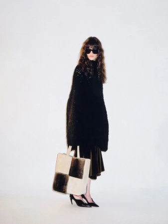Fashion model wearing an oversized black knit top, dark sunglasses, and holding a checkered shearling tote bag against a white backdrop.
