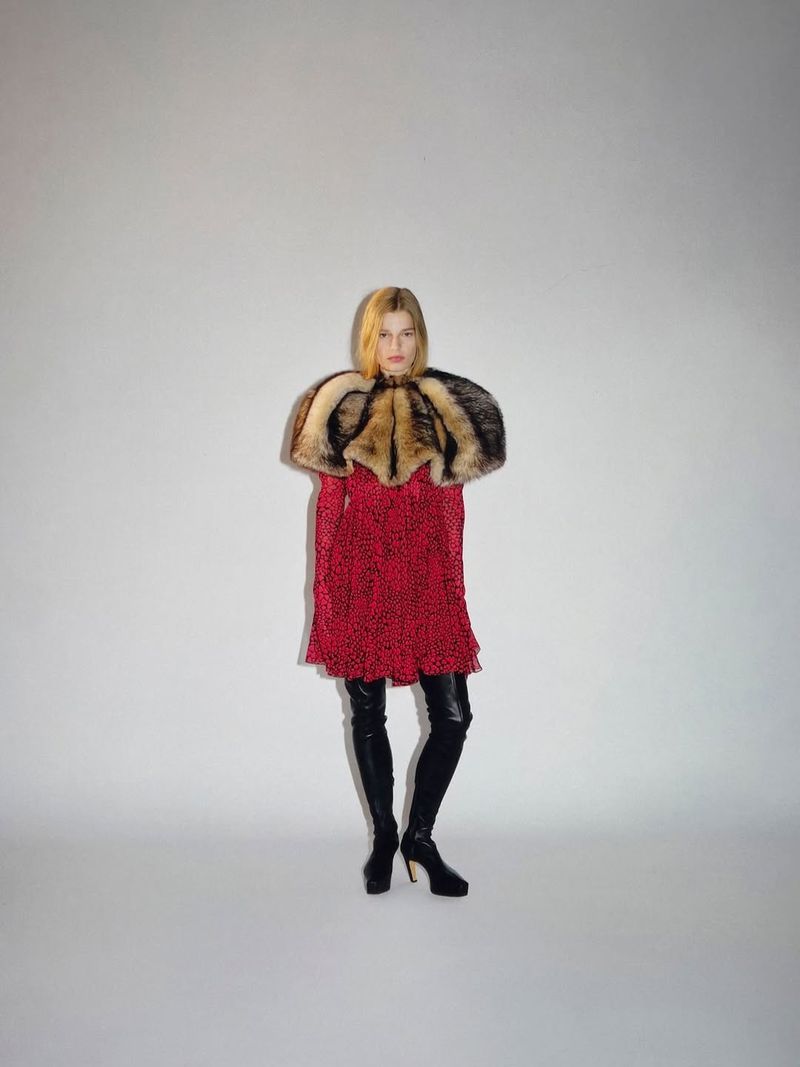 Model posing in a red patterned mini dress with a voluminous fur shoulder cape and black over-the-knee leather boots against a grey studio backdrop.