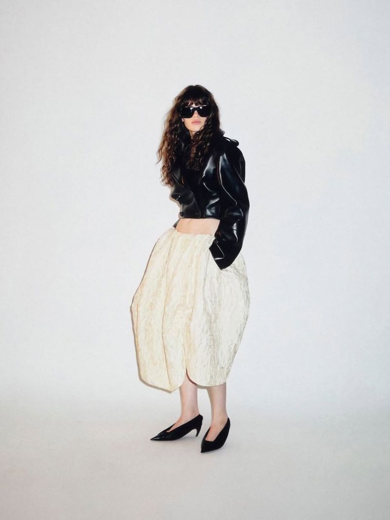Model wearing a black leather cropped jacket and cream skirt against a seamless white studio background.