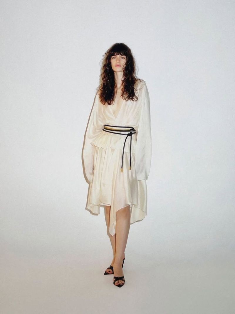 Model wearing an ivory draped dress with a black cord belt and strappy heels against a white studio background.