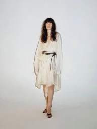Model wearing an ivory draped dress with a black cord belt and strappy heels against a white studio background.