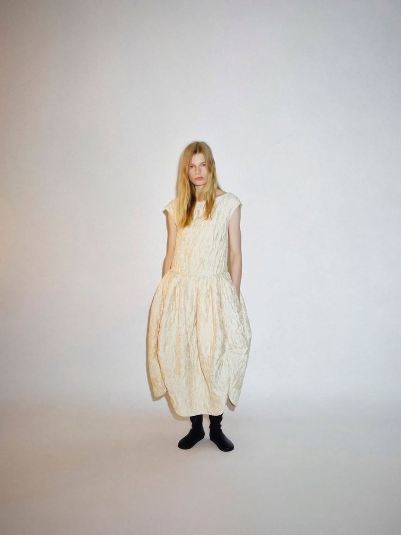 Model standing in an ivory textured sleeveless balloon dress with black shoes against a white studio backdrop.