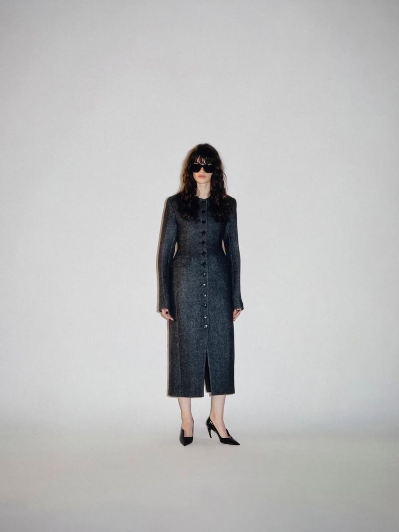 Model wearing dark sunglasses and a long charcoal button-front tailored coat with black pointed heels on a white studio backdrop.