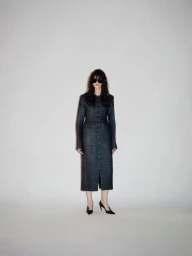 Model wearing dark sunglasses and a long charcoal button-front tailored coat with black pointed heels on a white studio backdrop.