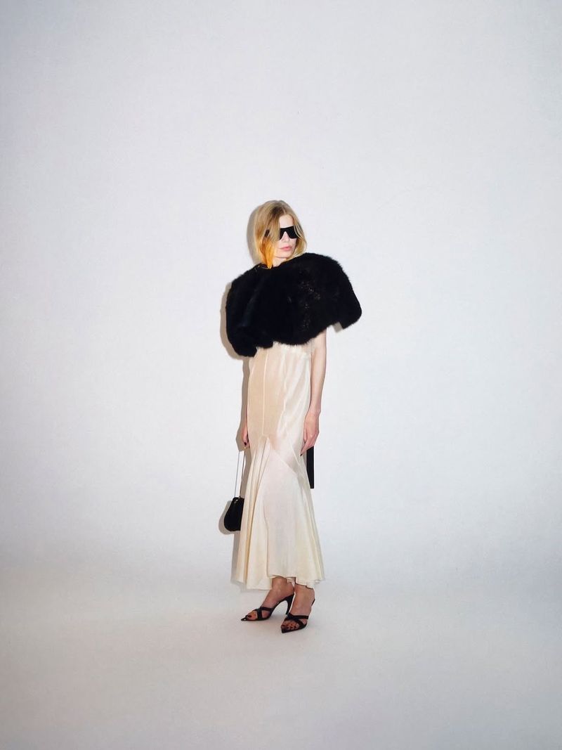 Model wearing dark sunglasses, a voluminous black textured shrug, and a long cream satin dress against a white studio backdrop.