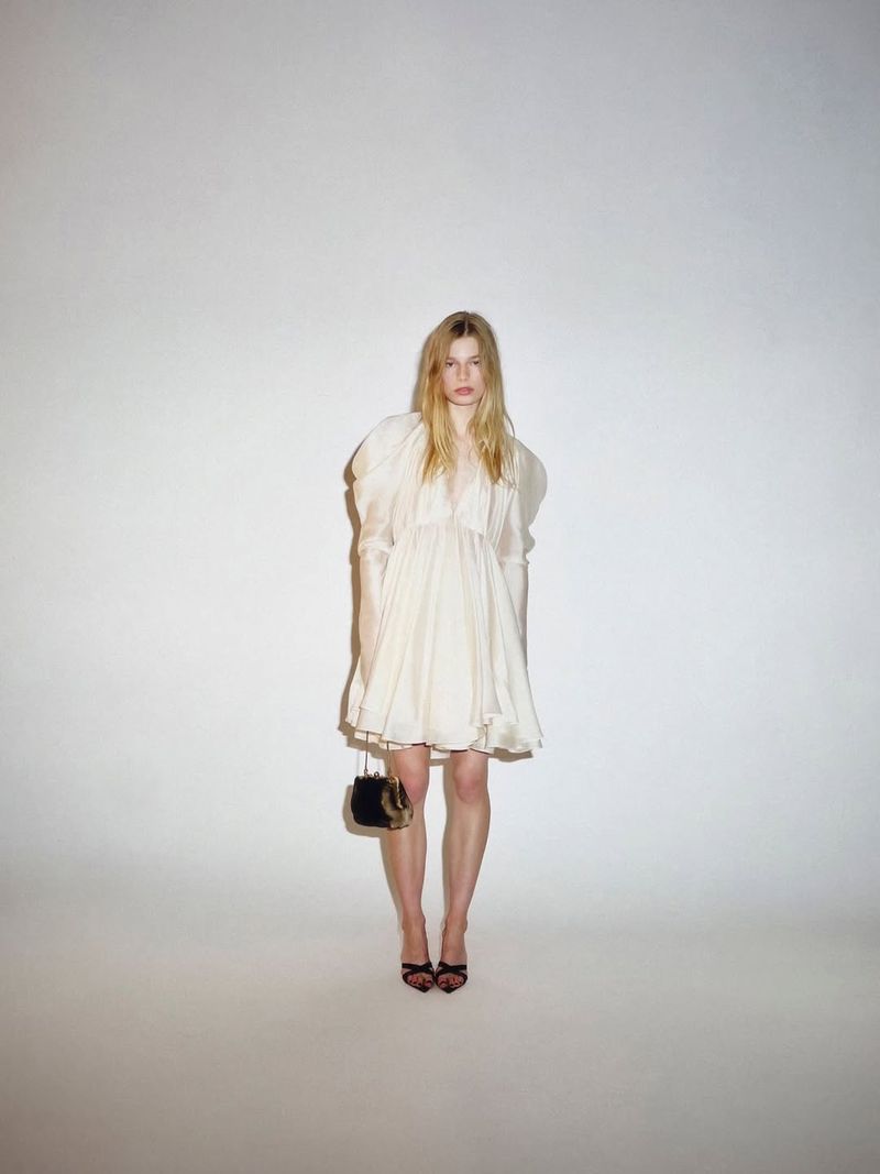 Model standing against a white studio background wearing an off-white voluminous mini dress, black heels, and holding a small handbag.