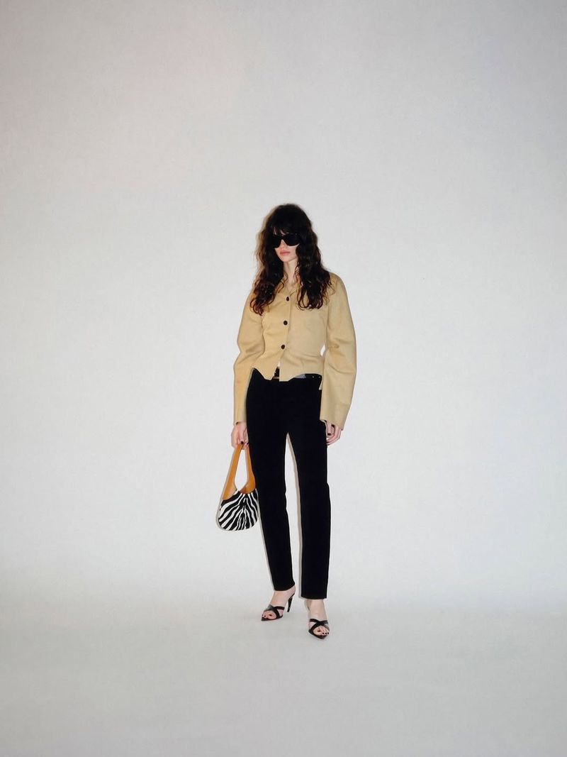 Model wearing dark sunglasses, a structured beige jacket, black trousers, and strappy heels holding a zebra print bag on a white backdrop.