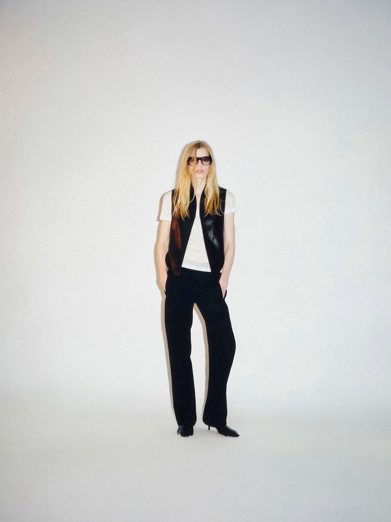 Model wearing dark sunglasses, a black sleeveless vest over a white t-shirt, and black trousers against a white seamless background.