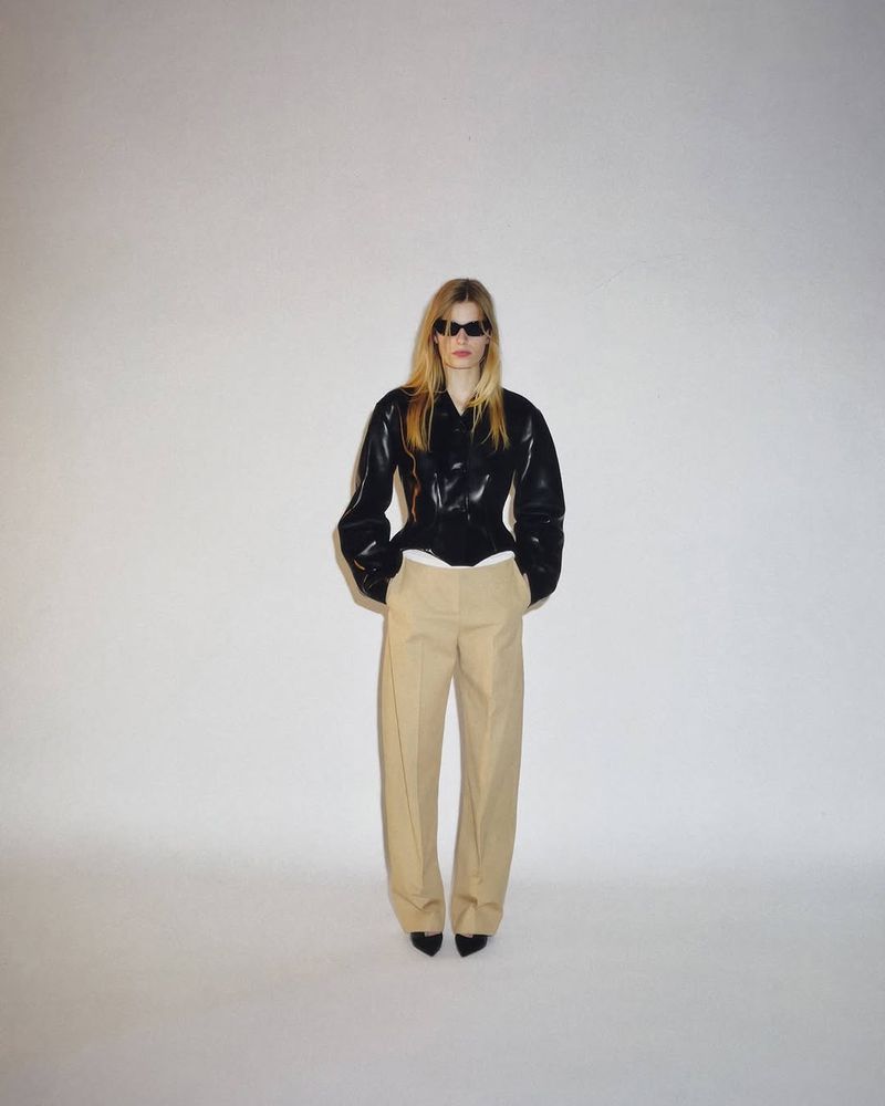 Model wearing dark sunglasses, a glossy black structured jacket, and beige tailored trousers against a white studio background.