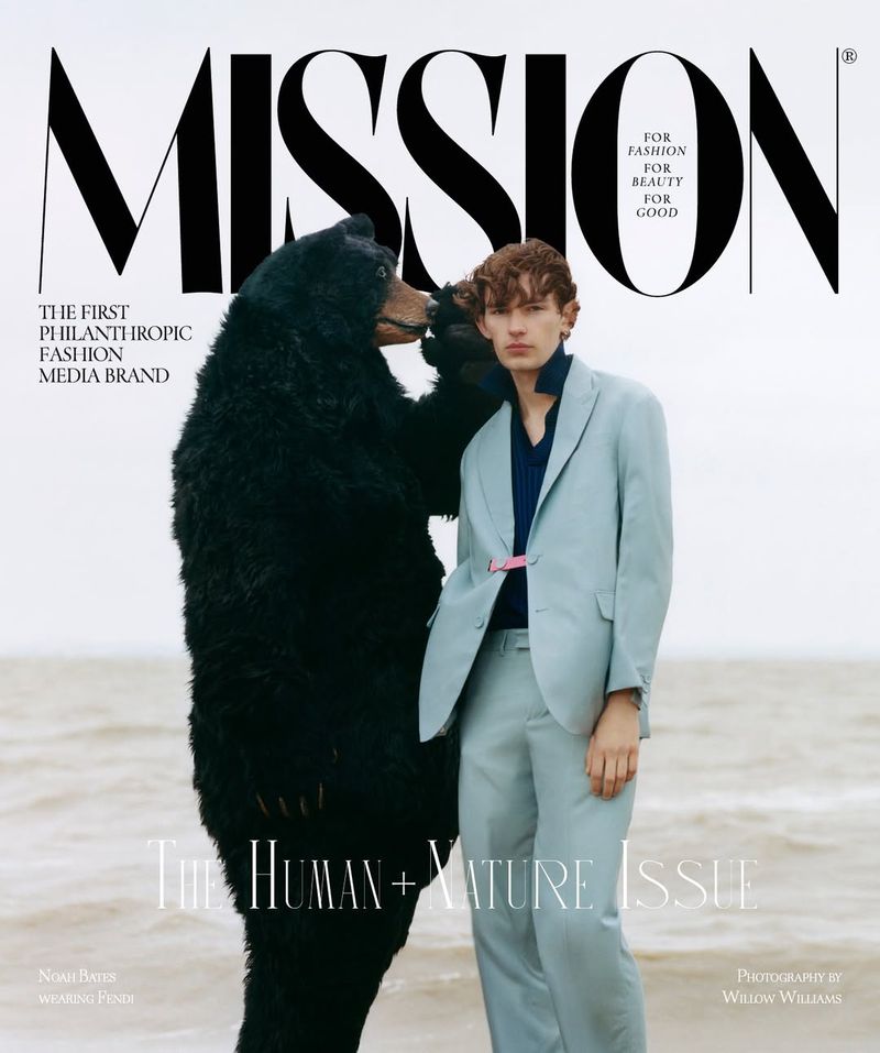 Model Noah Bates in a light blue suit posing next to a black bear costume on an overcast beach for Mission Magazine.
