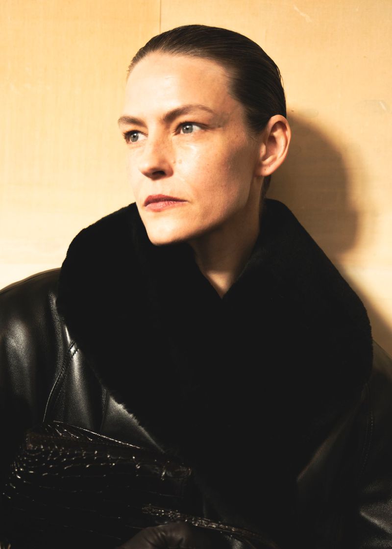 Model Malgosia Bela wearing a black leather coat with an oversized plush collar against a warm neutral backdrop.