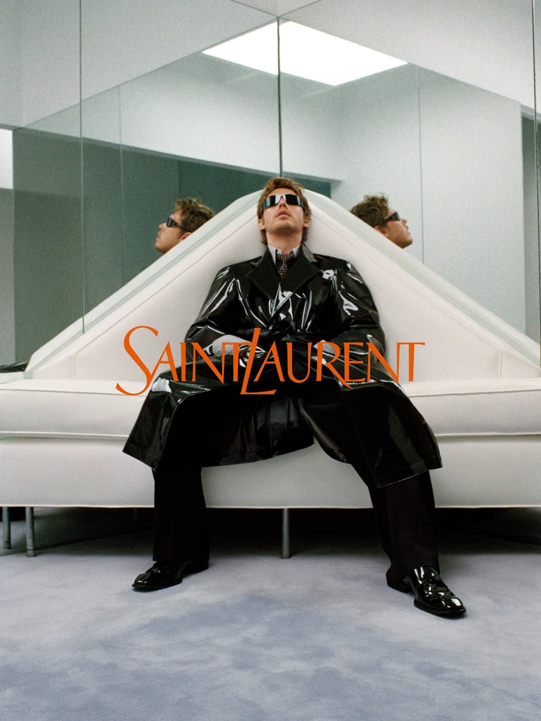 Austin Butler in a glossy black vinyl trench coat and sunglasses seated on a white sofa in front of mirrors for Saint Laurent.