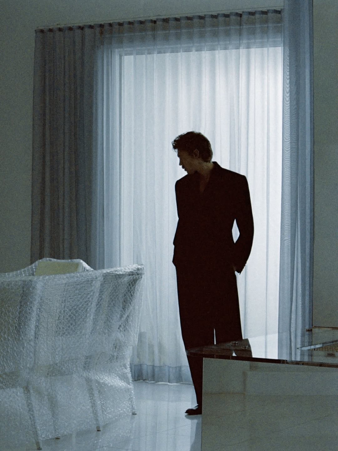 Austin Butler wearing a dark tailored suit stands in profile before sheer white curtains beside bubble-wrapped dining chairs.