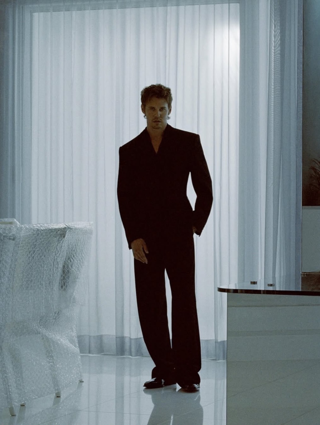 Austin Butler standing in a tailored black suit indoors in front of white sheer curtains with bubble-wrapped chairs nearby.
