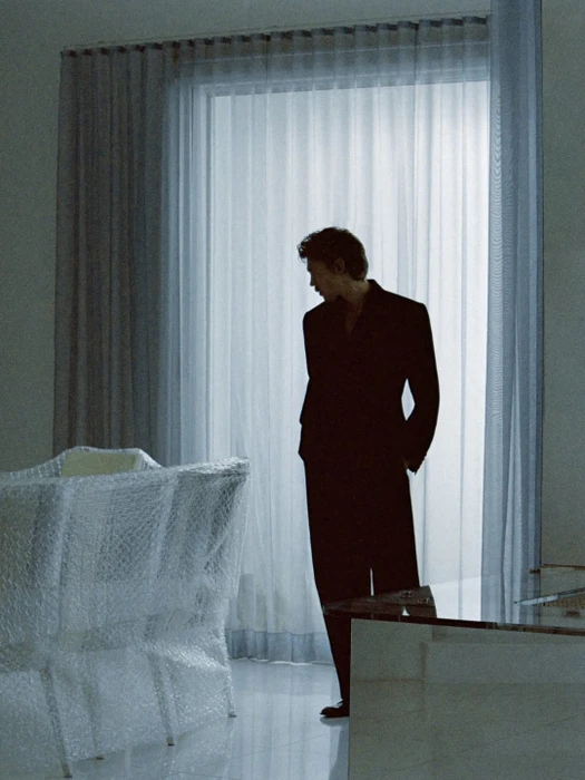 Austin Butler in a black suit standing beside sheer draped curtains near bubble-wrap covered chairs.