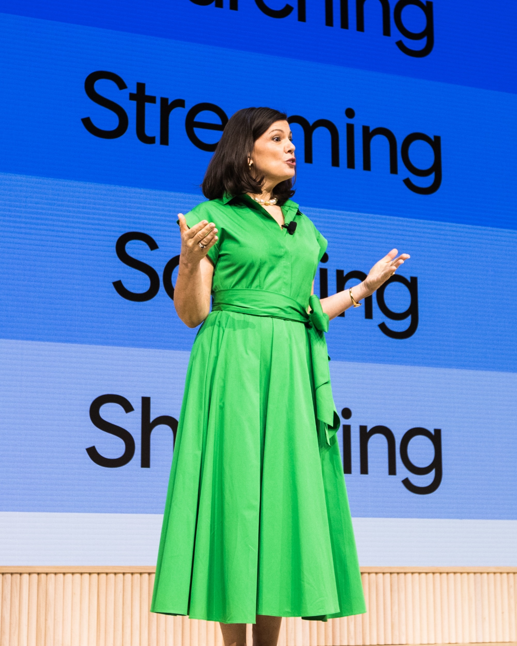 A woman wearing a vibrant green collared shirt dress speaking on stage in front of a blue typographic presentation screen.
