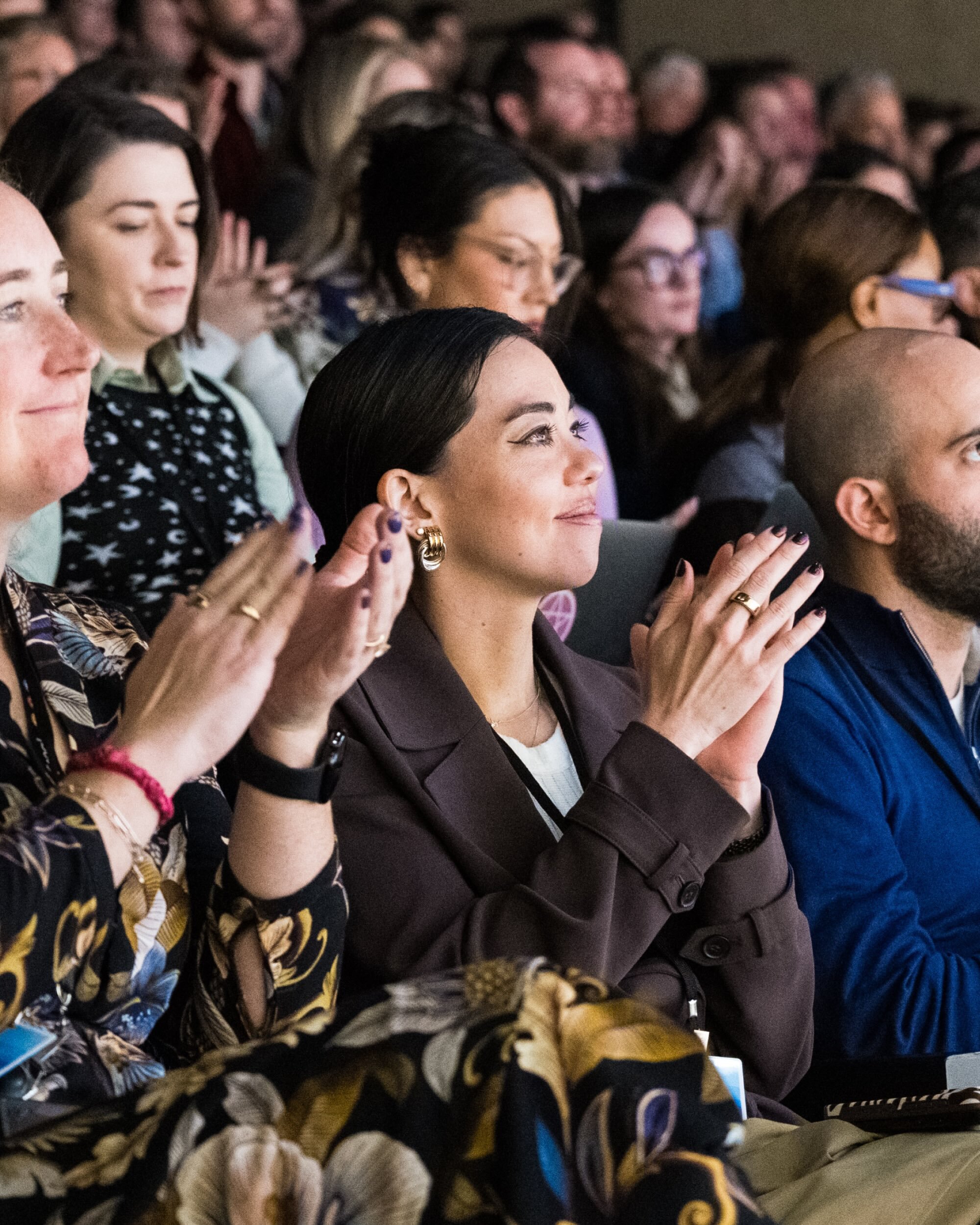 Woman in a brown blazer clapping alongside an attentive audience at an indoor conference event.