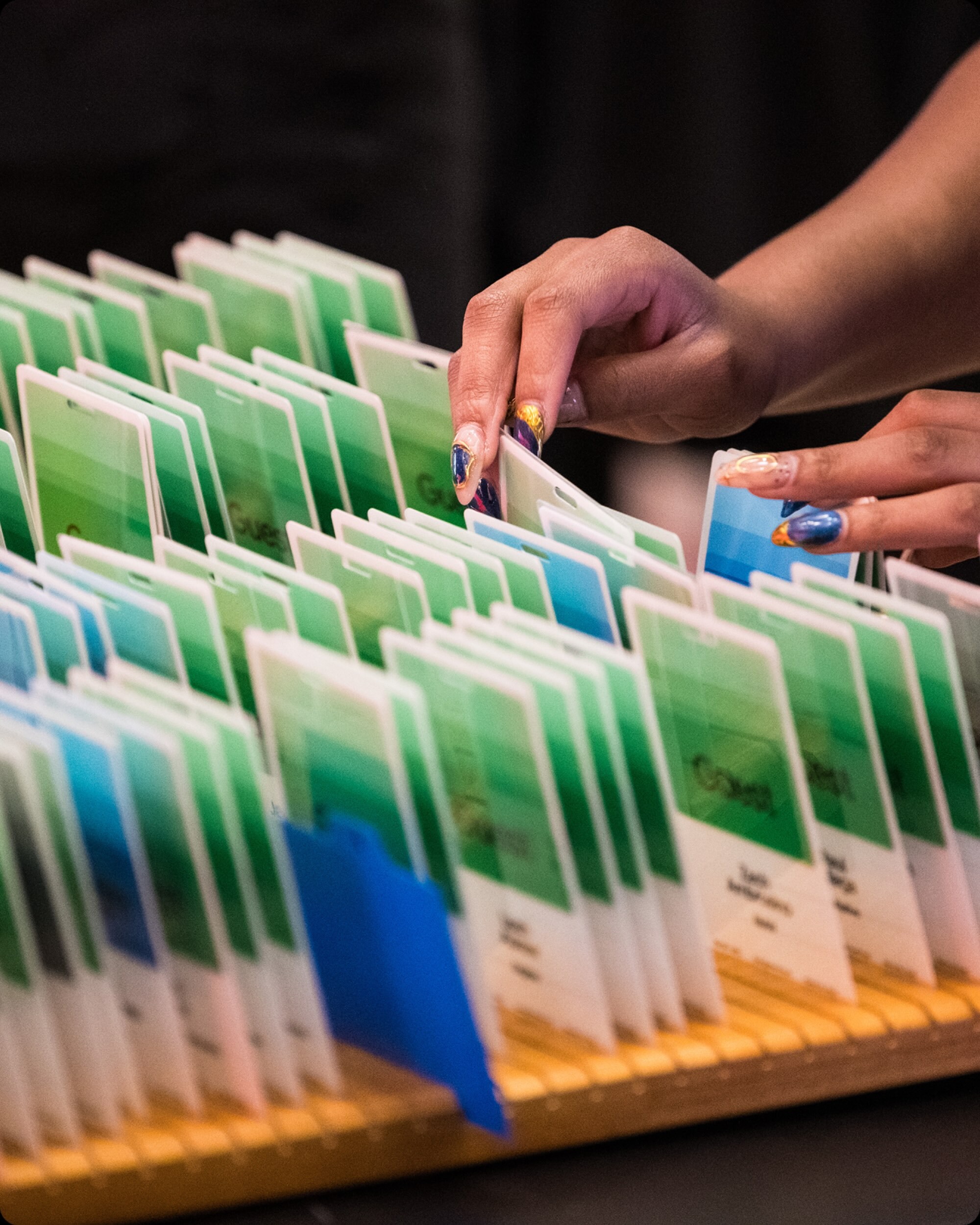 Close-up of manicured hands sorting through printed green and blue gradient guest name badges in a slotted wooden organizer.