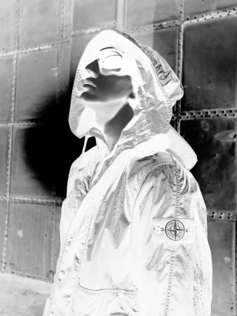 Monochrome high-contrast portrait of a person wearing a hooded technical jacket and sunglasses against a riveted metal wall.