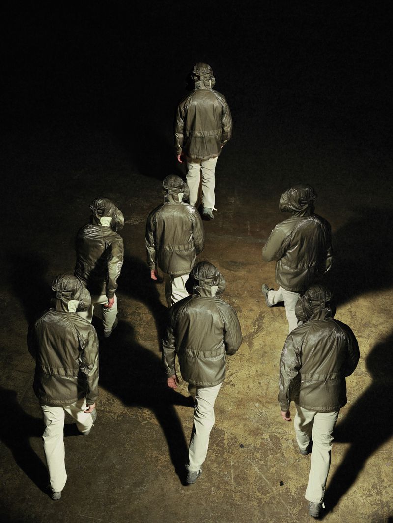 High-angle view of models wearing matching olive hooded jackets and beige trousers walking across a dark surface under a spotlight.