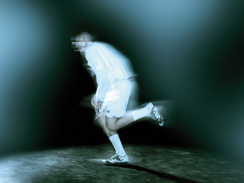 Motion-blurred man running in a white athletic kit and football boots against a dark, cool-toned illuminated ground.