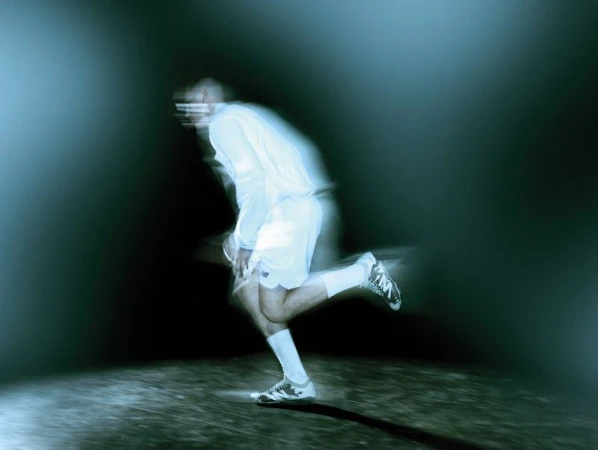 Motion-blurred man running in a white athletic kit and football boots against a dark, cool-toned illuminated ground.