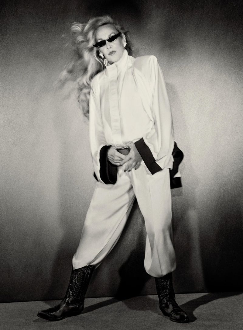 Model Jerry Hall poses in a white draped suit, dark sunglasses, and black cowboy boots against a neutral studio backdrop.