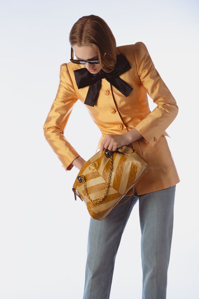 Model Jess PW wearing a gold tailored jacket with a black bow while looking into a tan patterned handbag on a white background.