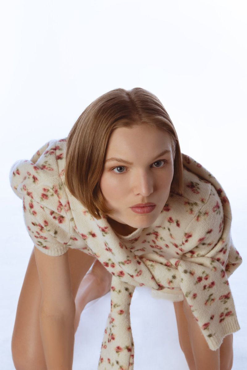 Model with brown hair wearing a floral knit sweater and scarf against a white background