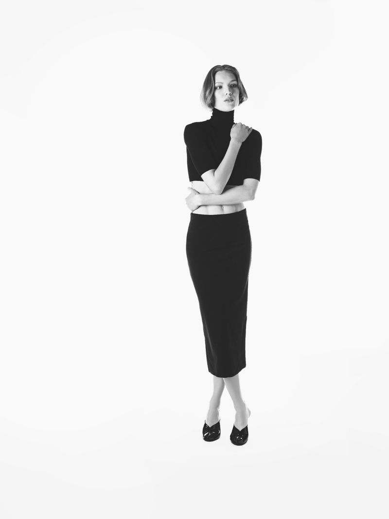 Model Jess PW posing in a black cropped turtleneck and black midi skirt against a solid white studio background.