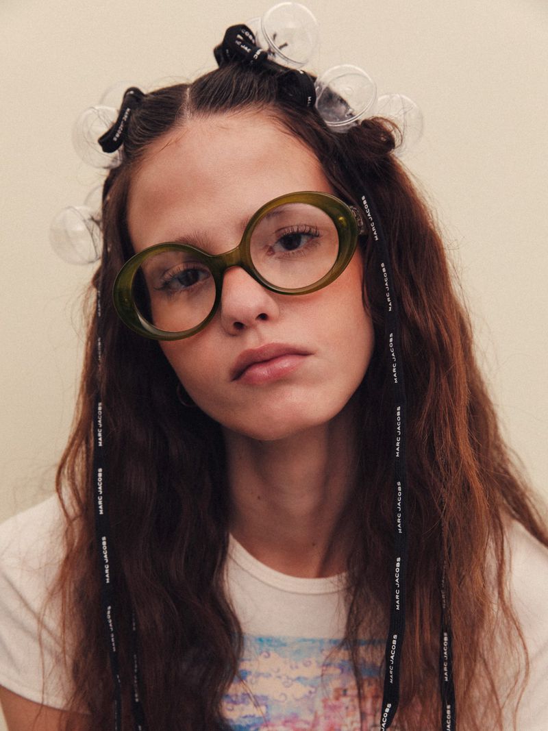 Model wearing round olive green glasses and clear spherical hair rollers tied with black ribbons against a seamless backdrop.