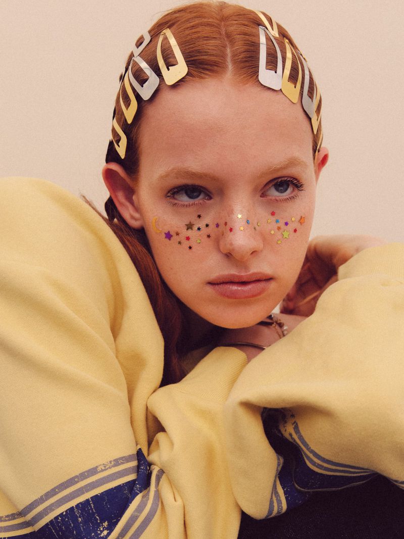 Model with red hair wearing gold and silver snap clips, star freckle makeup, and a yellow sweatshirt against a pale background.