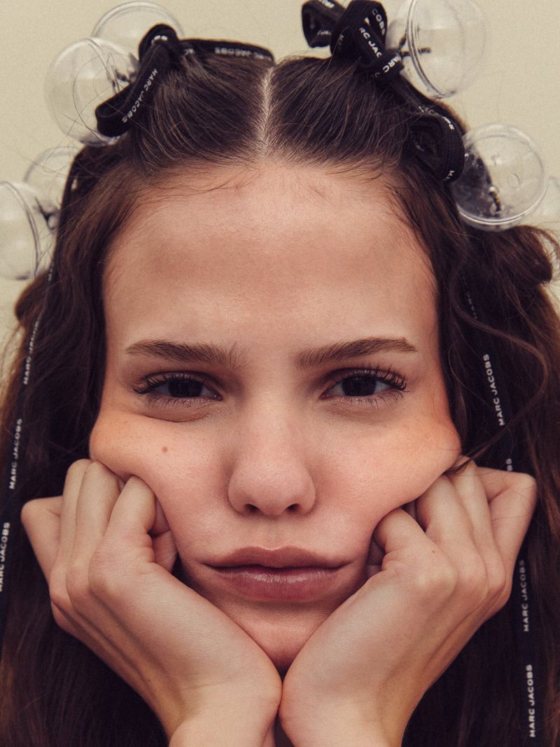 Close-up portrait of a woman resting her face on her hands with Marc Jacobs branded ribbon hair accessories on a neutral background.