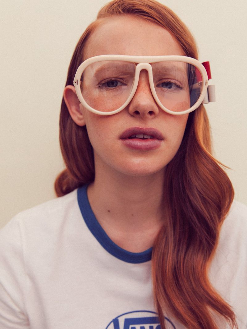 Close-up portrait of a red-haired woman wearing oversized white framed glasses and a white ringer t-shirt.