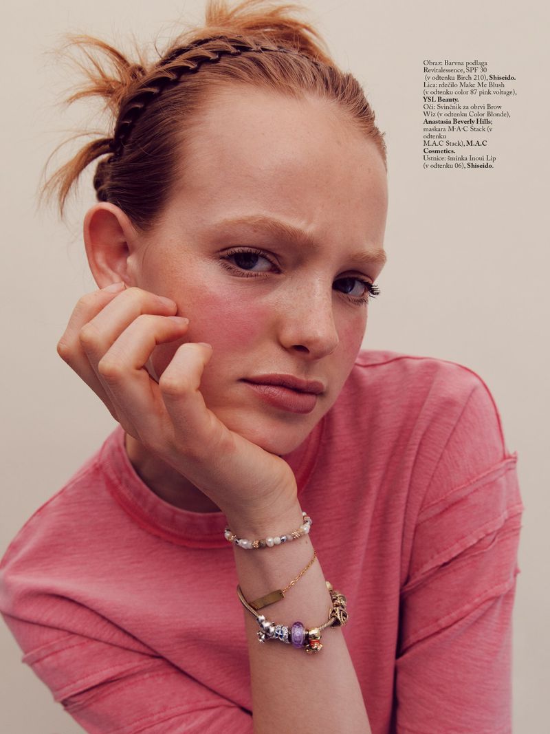 Close-up portrait of a young woman resting her chin on her hand, wearing a pink t-shirt and layered bracelets.