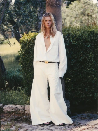 Model Freya Nutter standing outdoors leaning against a pillar in an open white shirt and wide-leg trousers.