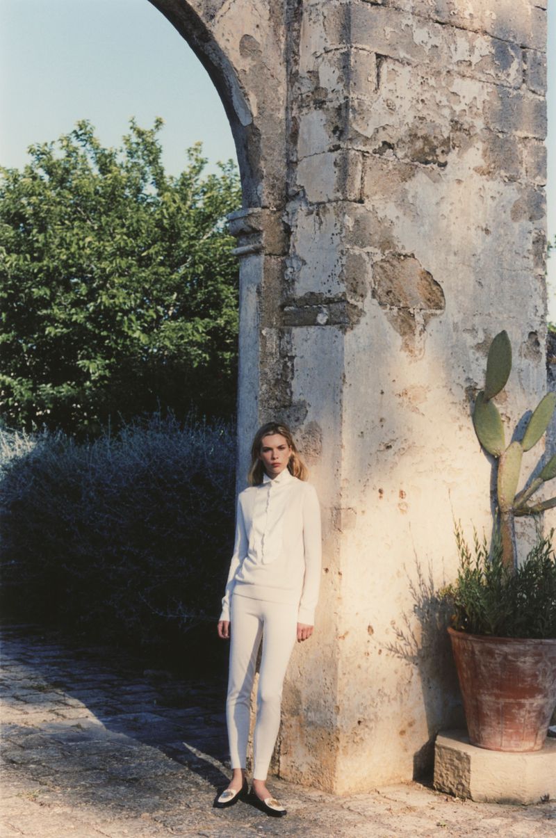 Model Freya Nutter poses in an all-white top and trousers while leaning against a rustic stone arch beside a potted cactus.