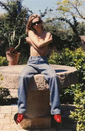 Model Freya Nutter wearing blue wide-leg trousers and dark sunglasses, seated outdoors on a stone pedestal in bright sunlight.