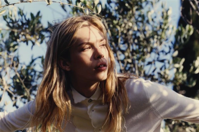 Model Freya Nutter in a cream collared shirt with dappled sunlight on her face against leafy olive branches.