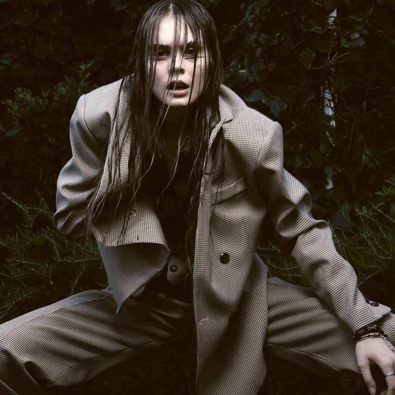 An adult female crouches in an outdoor setting, wearing a tailored suit with wet hair, in a fashion-forward shot.