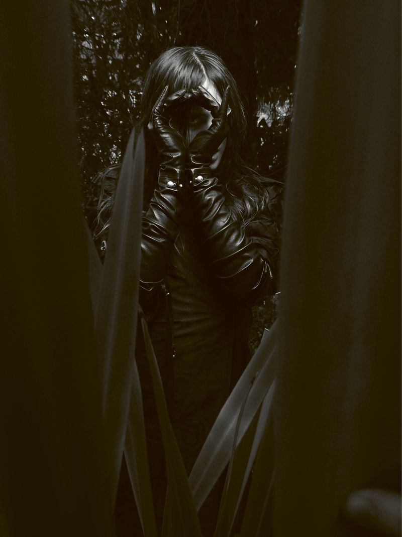 Person wearing black leather gloves peering through hands around eyes, framed by foreground leaves in a dark outdoor setting.