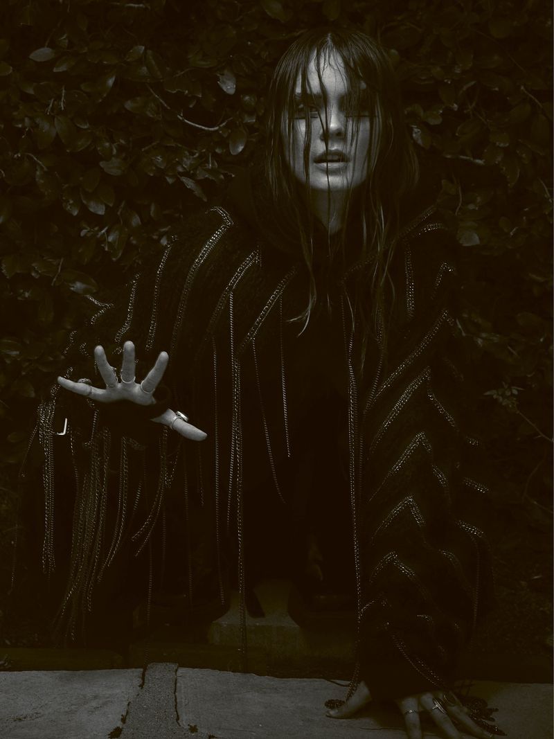 Woman with dark eye makeup and fringed poncho poses outdoors against a leafy green background in a fashion editorial style photograph.