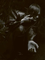 Woman in a dark leather jacket posing outdoors among dense foliage in low-key lighting for The Hunger Magazine.