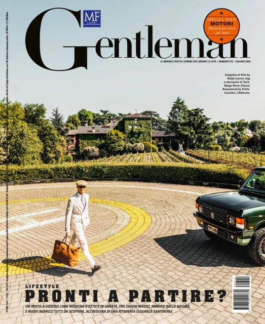 Man in a white suit holding a brown leather bag walking past a green vintage SUV on a paved driveway