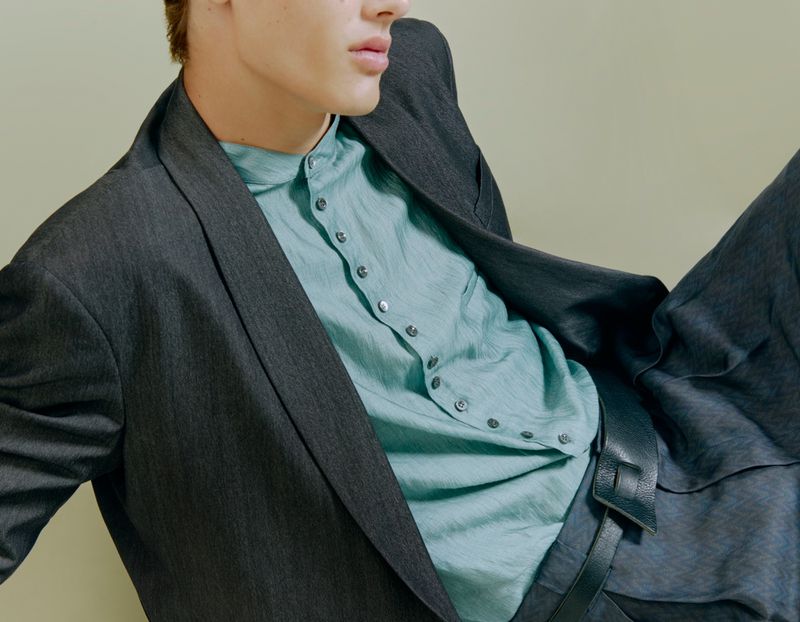 Close-up portrait of a male model wearing a grey blazer, turquoise button-up shirt, and dark trousers against a plain wall.